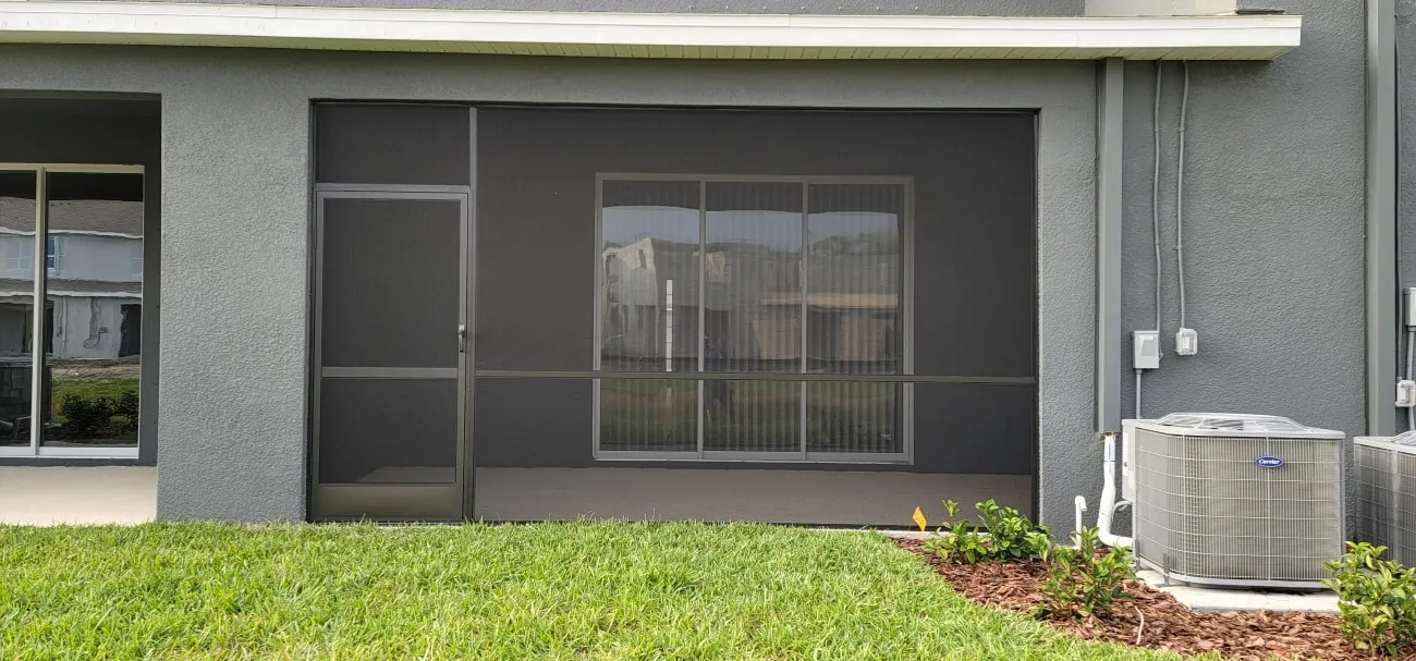 Screened porch with aluminum door on a dark gray Florida home beside landscaped beds and AC units