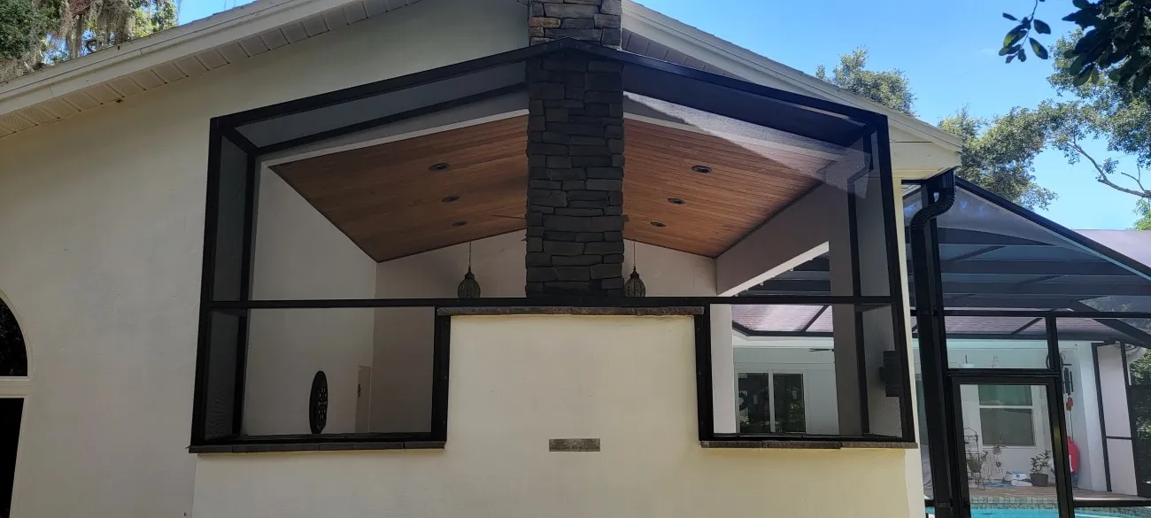 Screen enclosure over an outdoor living area with vaulted wood ceiling and stone chimney, pool cage beyond