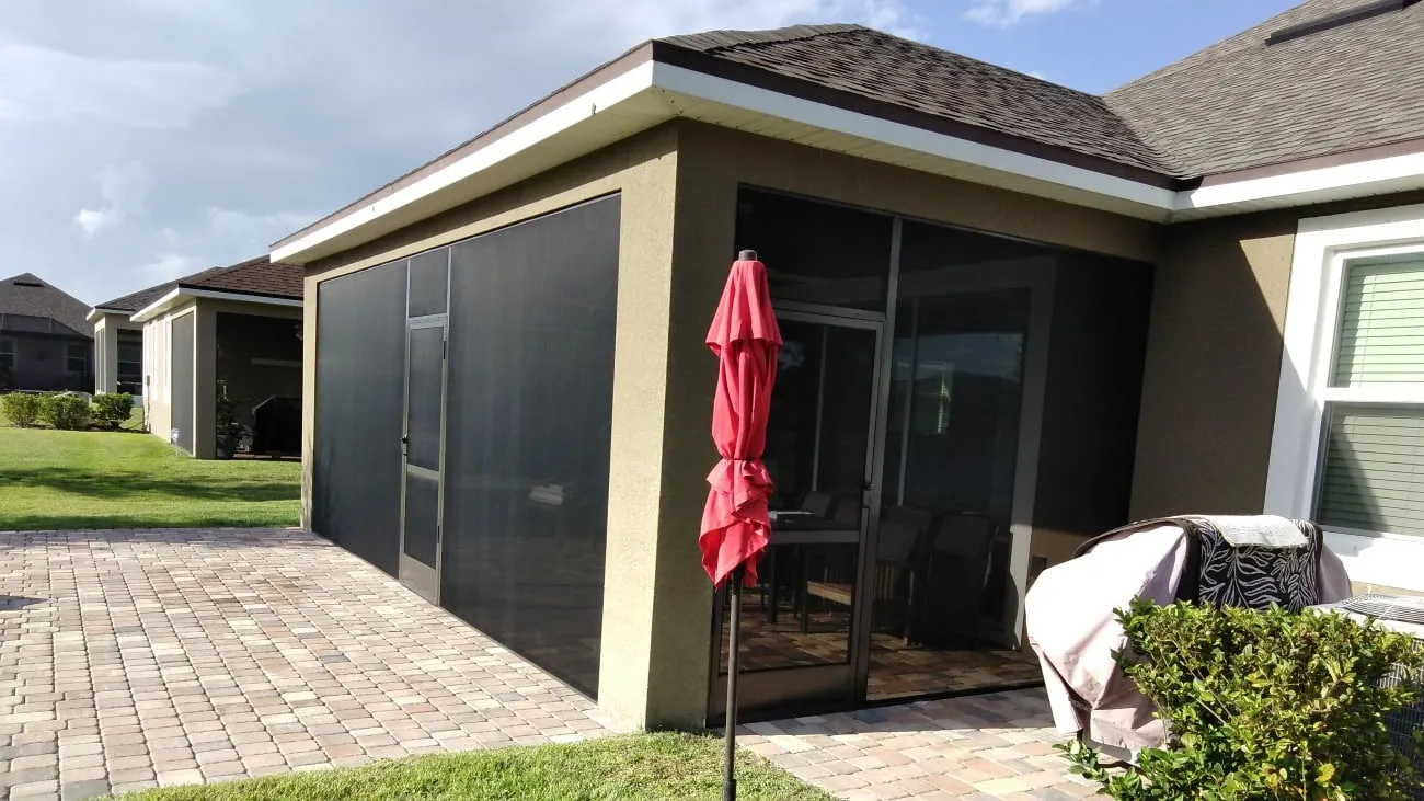 Corner screened lanai with screen door on a tan Florida home, framed by a brick paver patio and red umbrella
