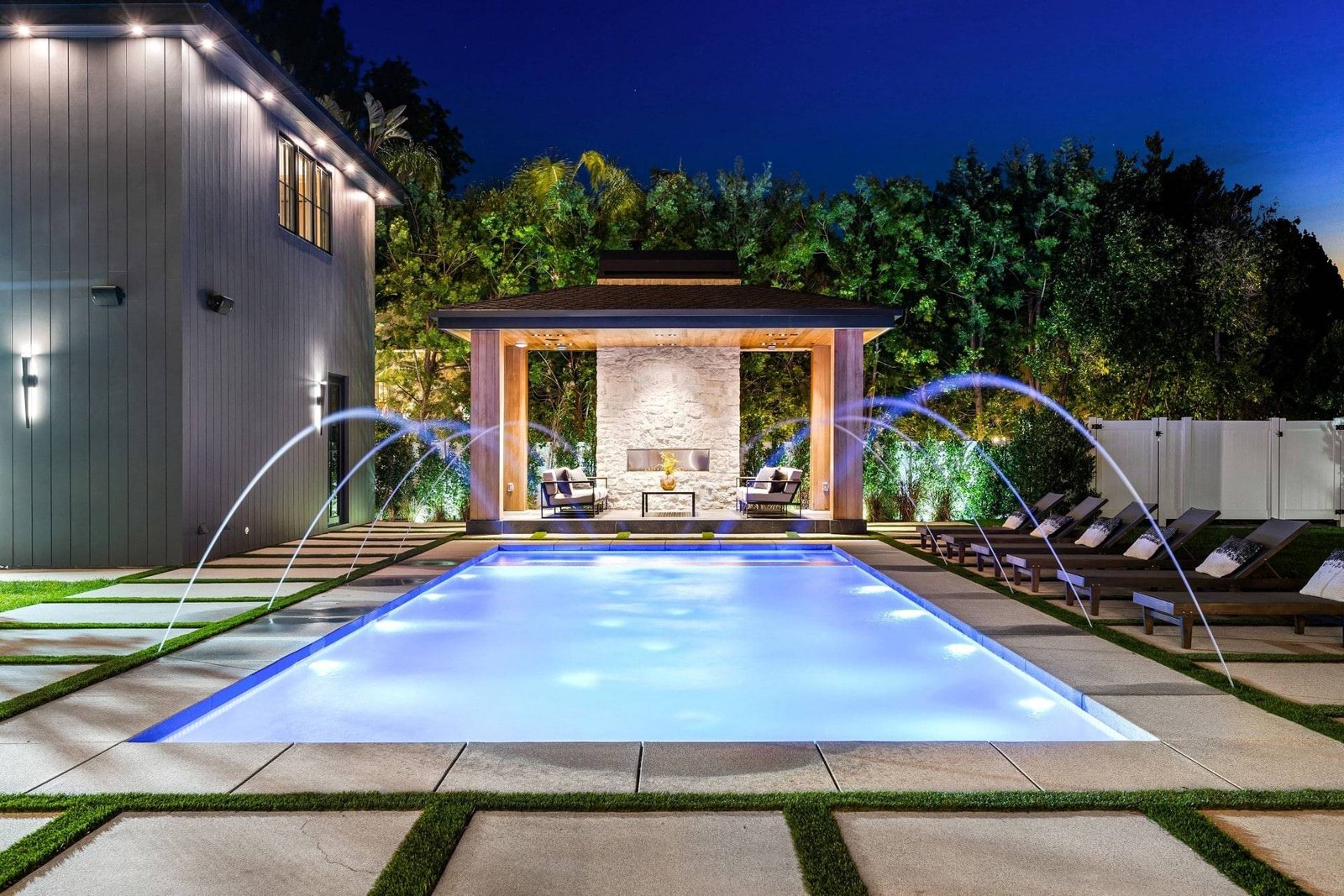 Rectangular pool lit at night with arcing deck jets and a stone fireplace cabana behind a modern Florida home