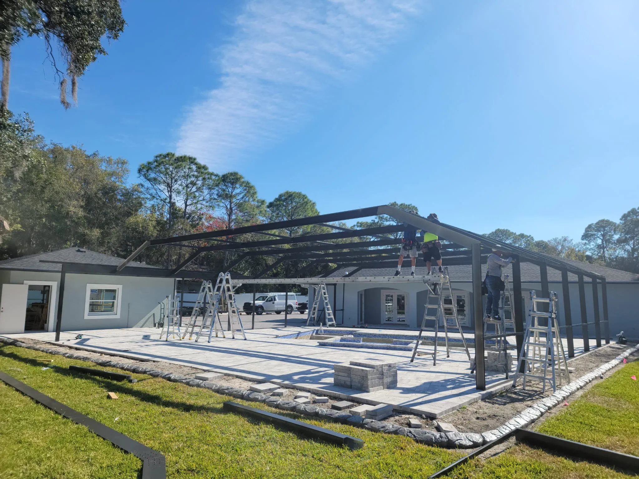 Crew on ladders raising the gable roof trusses of an aluminum pool cage over a new pool deck