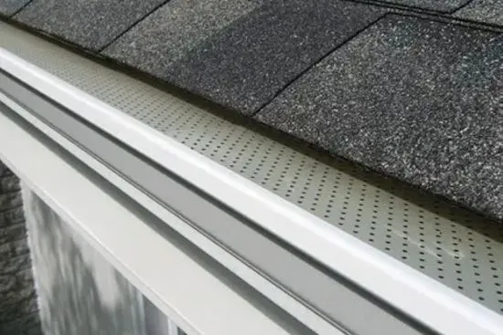 Close-up of a white aluminum gutter with a perforated leaf guard installed beneath a dark shingle roof