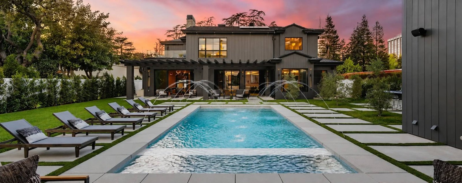 Modern backyard pool with arcing deck jets and lounge chairs in front of a dark two-story home at sunset