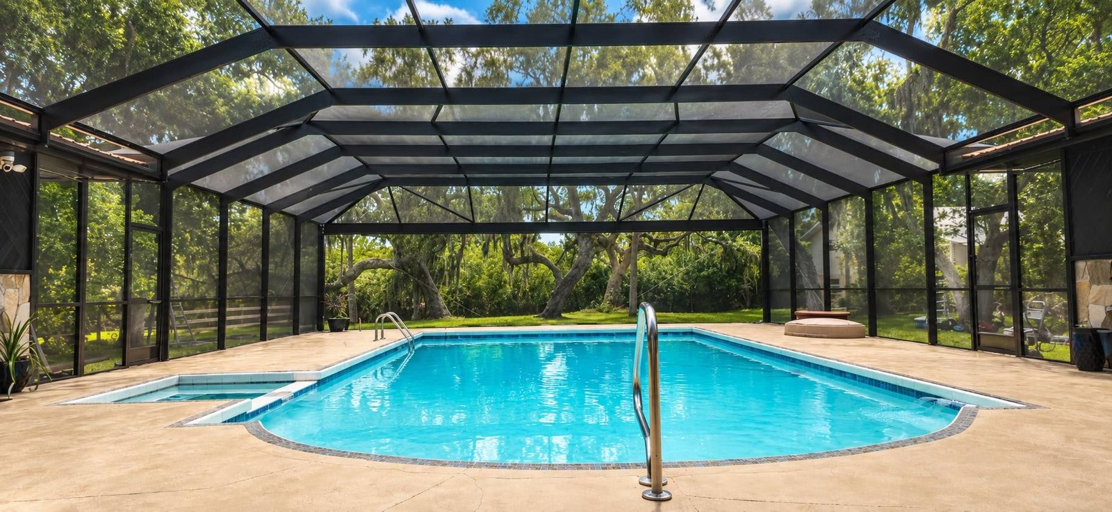Inside a panoramic pool enclosure with wide dark aluminum spans and few uprights, looking out over the pool to the oaks beyond