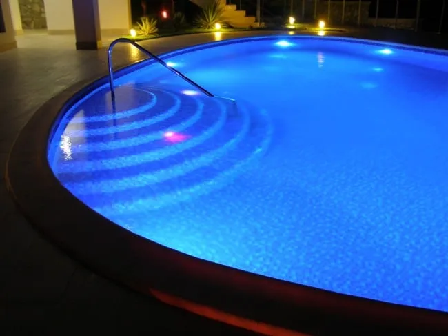 Curved pool glowing bright blue at night from underwater LED lights, with landscape lighting along the deck