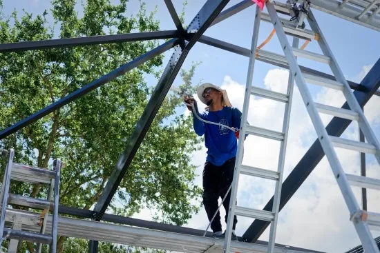 Worker on a ladder spray-painting the roof beams of an aluminum pool cage
