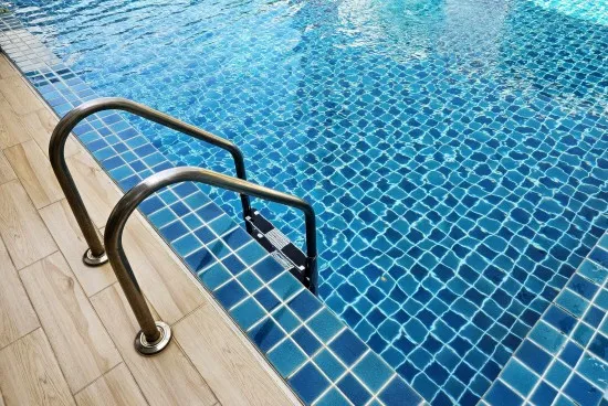 Stainless steel handrail on pool steps with a shimmering blue tile finish beside a wood-look paver deck
