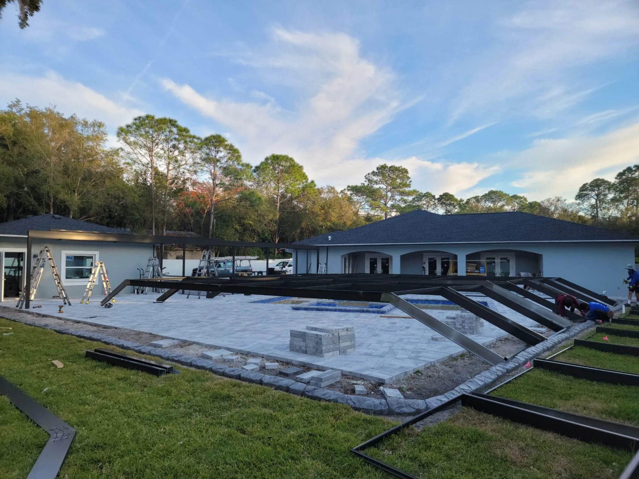 Aluminum frame sections staged on the paver deck around an unfinished pool at the start of a pool cage build