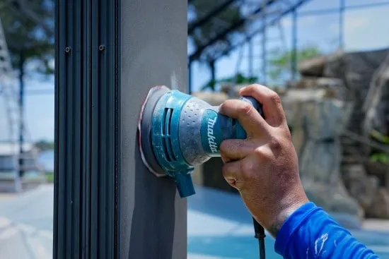 Worker sanding an aluminum pool cage column with an orbital sander to prep the surface for paint