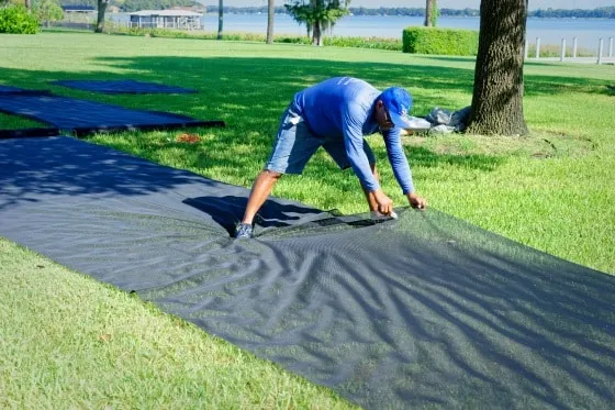 Technician laying out and cutting black screen mesh panels on a lakeside lawn before installation