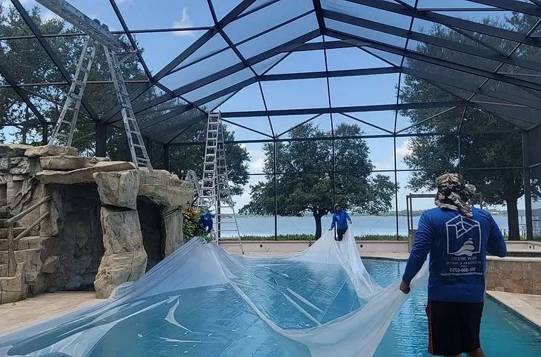 Crew covering a lakefront pool with protective plastic sheeting before cage restoration work begins