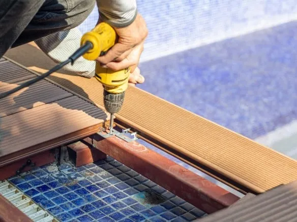 Worker using a cordless drill to fasten composite deck boards beside a blue mosaic-tiled pool