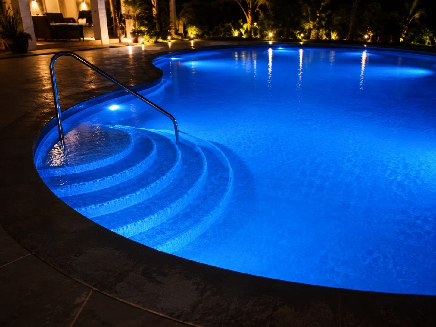 Night photo of a curved swimming pool with blue LED lighting and submerged steps leading into the water.