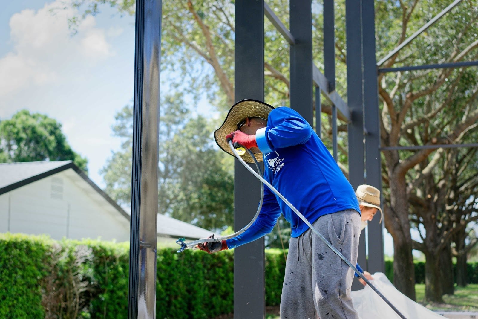 Worker in blue shirt and straw hat spray painting a dark metal frame or structural element outdoors.